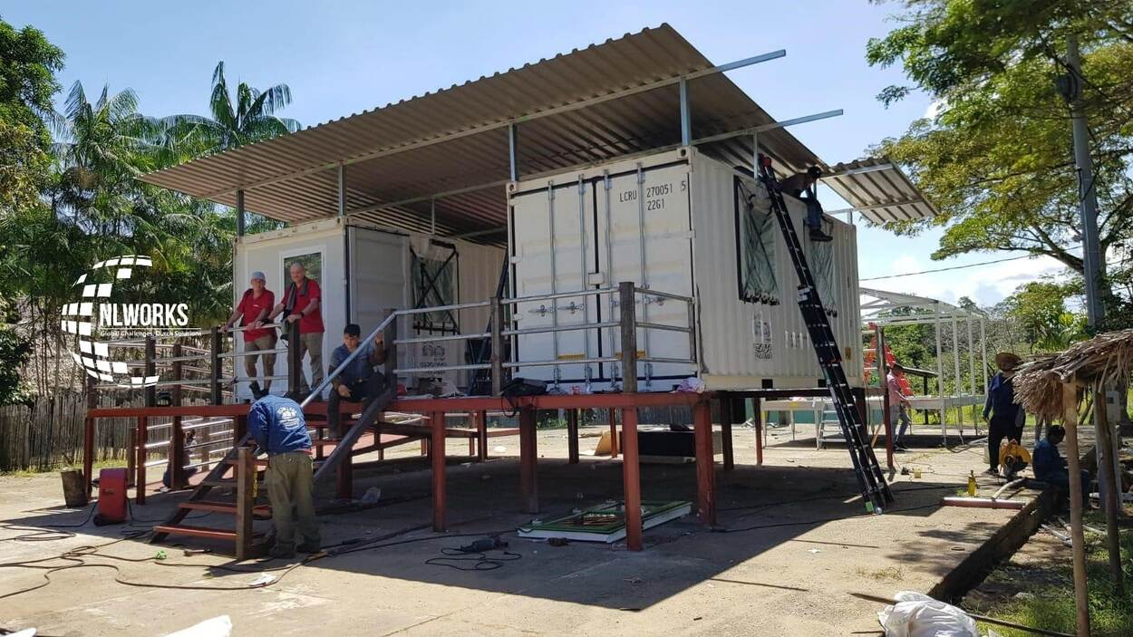 People stand on the balcony of a stilted and under construction house in Colombia. There is greenery and a countryside like atmosphere in the background.