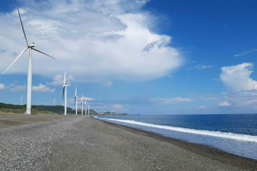 A shoreline with windmills. The sand meets the ocean and blue skies and clouds are seen in the background.