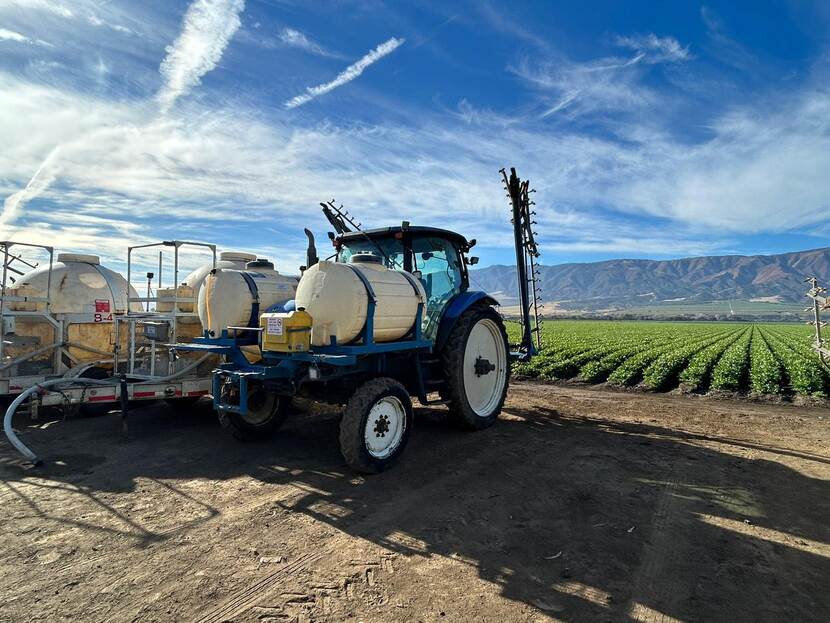 A tractor with tanks on it stands next to a Califronian field. Behind the tractor is a lush green field with rows of agriculture and mountains in the distance.