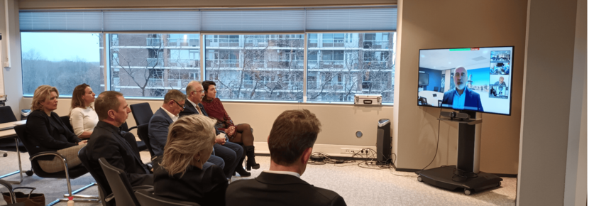 Partners gathered in a conference room watching the screen where a man is on video conference with the room