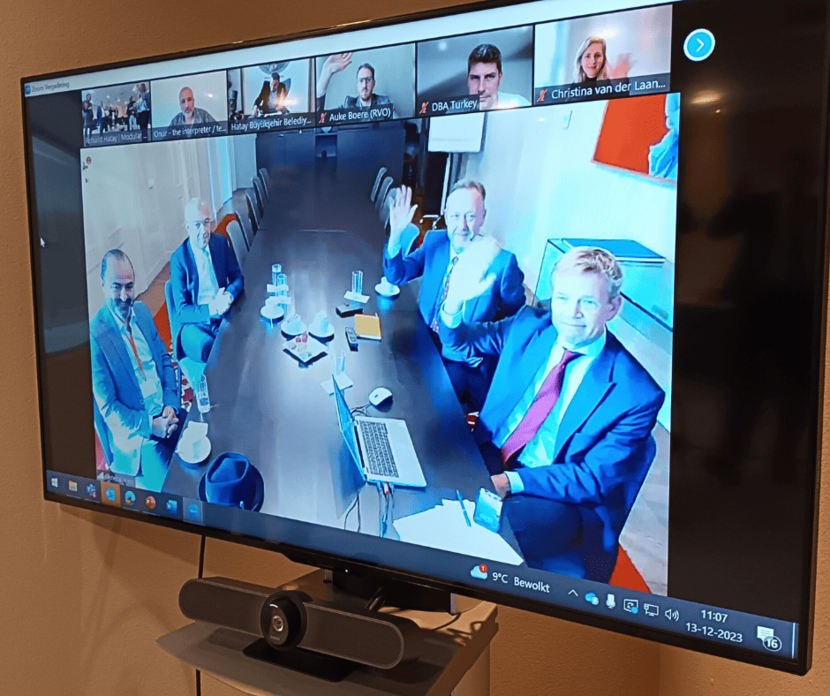 A screen displays partners in Turkey seated at a table in suits and waving while smiling at the camera