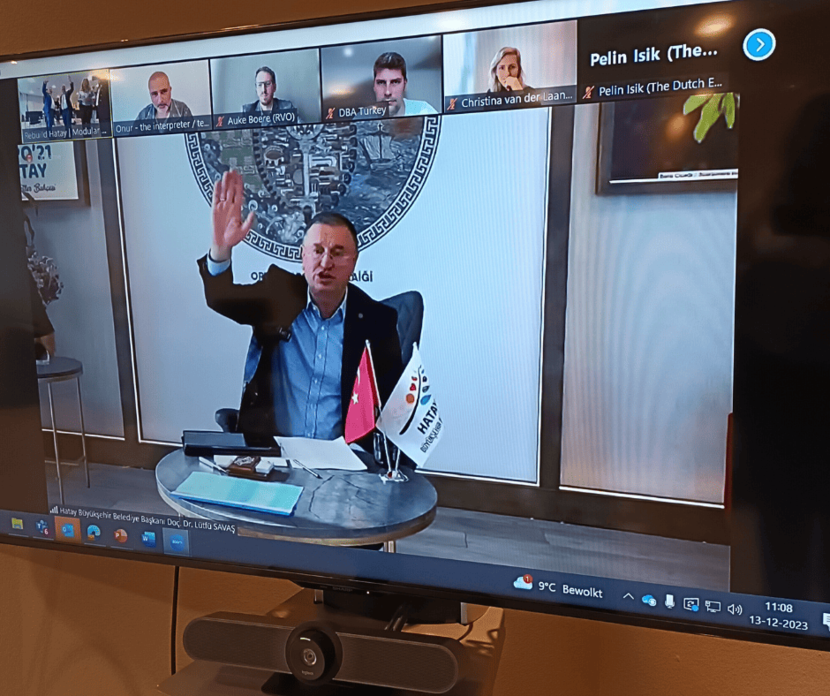 A screen displays a video conference showing a Turkish partners seated at his desk with a Turkish flag on the desk waving at the camera and smiling