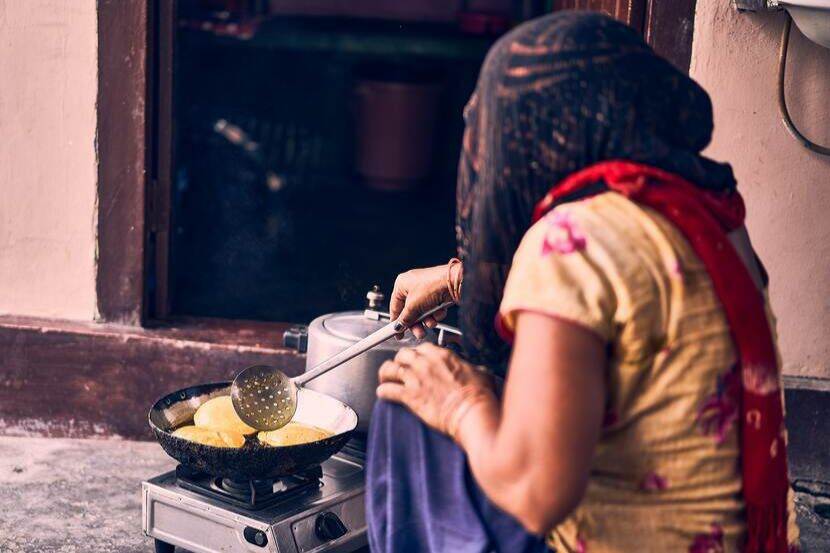 A lady in a headscarf is frying dough in a wok that is on a stove on the floo of the outside of a mud home